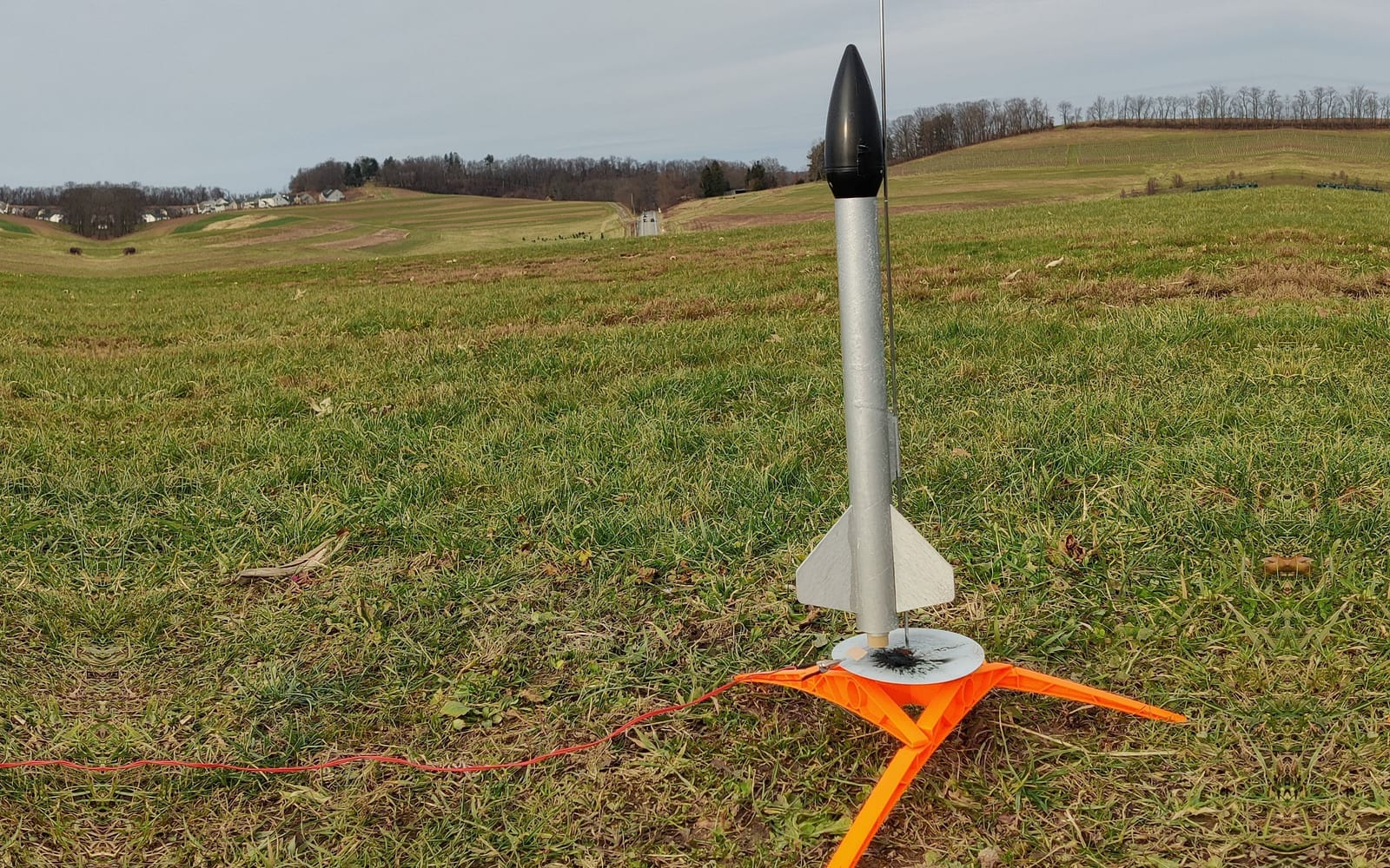 A silver model rocket with a black nose cone standing on an orange 3D-printed launch pad in a grassy field